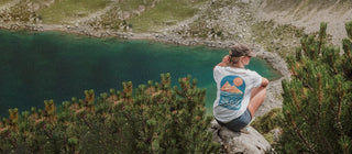 Eine Junge Frau sitz in den Alpen auf einem Fels beim Wandern. Sie trägt ein Weißes T-Shirt der Marke The Clue Clothing. Man sieht einen Bunten Backprint. Das Shirt ist ein faires T-Shirt in Weiß mit buntem Rückendruck. Unisex getragen von einer Damen. Das T-Shirt wird in Portugal produziert. Fair produziertes Shirt. Made in EU.